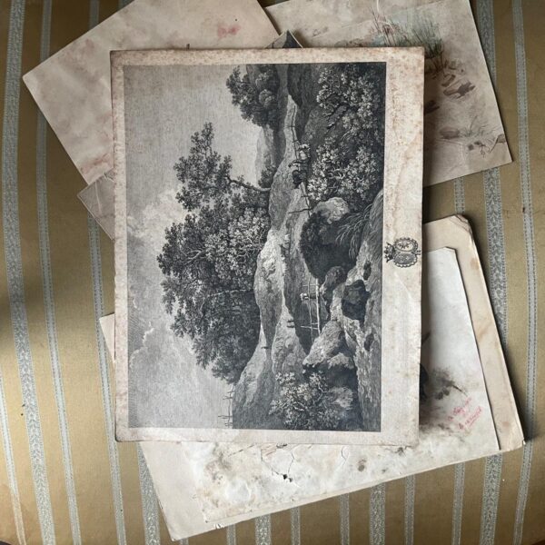 Gravure ancienne représentant un paysage rocheux, posée parmi d’autres estampes et aquarelles anciennes